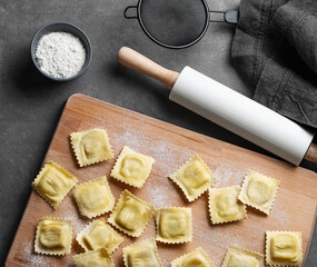 Homemade pasta ravioli on wooden cutting board on dark background with flour, napkin and rolling pin.