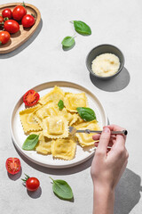 Hand holding fork in plate with homemade ravioli pasta with parmesan, tomatoes and basil on light background