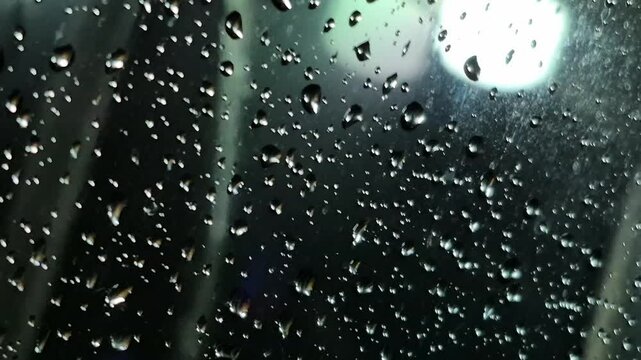 Rain at night on the side of the highway seen from the perspective of inside the car, you can see raindrops sticking to the car window. There is light from street lights, cars and other vehicles passi