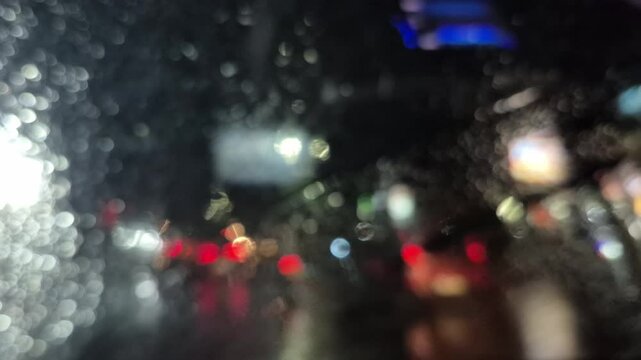 Rain at night on the side of the highway seen from the perspective of inside the car, you can see raindrops sticking to the car window. There is light from street lights, cars and other vehicles passi