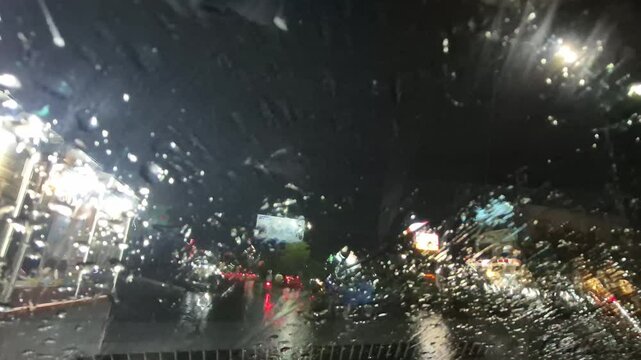 Rain at night on the side of the highway seen from the perspective of inside the car, you can see raindrops sticking to the car window. There is light from street lights, cars and other vehicles passi