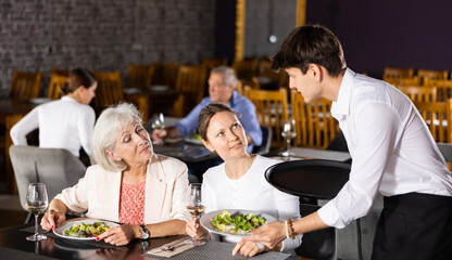 Amiable young waiter serving ordered dishes to carefree female friends gathered over dinner in restaurant