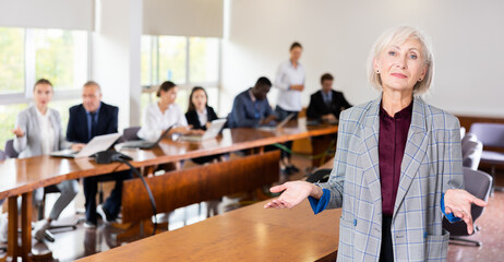 Portrait of positive senior businesswoman in suit gesturing during briefing in meeting room of company office.