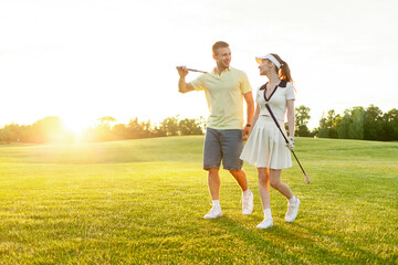 young sports couple in uniform playing golf together on field with lawn at sunset, woman with instructor practicing golf competitions and having active recreation