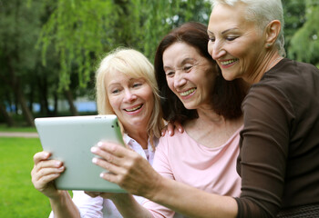 Obraz premium Three old women are smiling and looking photos at the at screen of the tablet pc in the park on a summer day. Friendship is priceless.