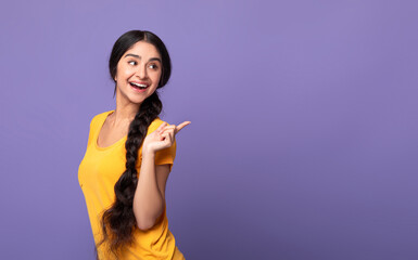 Obraz premium Special Offer. Portrait of smiling young indian woman with long braid pointing finger at copy space looking back isolated over purple studio background. Excited woman showing free space, place for ad