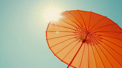 A vibrant orange umbrella with rays of sunlight shining through, set against the clear blue sky on sunny day background