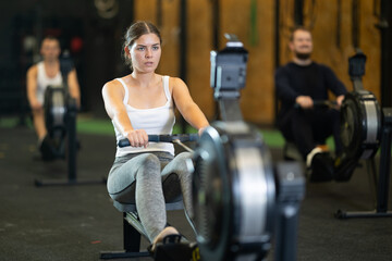 Determined young girl training with rowing machine in sports hall during crossfit workout