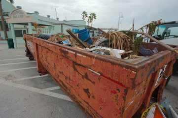 Roll-off Trash container and Green palm trees  in Parking  Lot Trash Beach damaged piled storm surge  after hurricanes Helene and Milton at Gulfport Beach in Florida. Debris, sand, orange sign and tem
