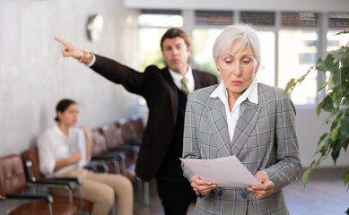 Portrait of upset senior businesswoman holding papers in hands while standing in office lobby with...