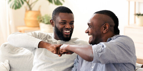 Father-Son Friendship. Black Millennial Guy Bumping Fists With His Senior Dad At Home, Enjoying Spending Time Together, Closeup