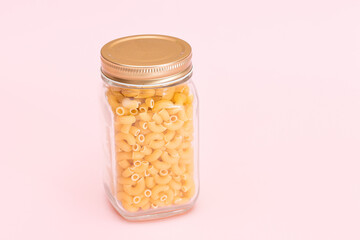 Uncooked Chifferi Rigati Pasta in Glass Jar on Pink Background. Fat and Unhealthy Food. Scattered Classic Dry Macaroni. Italian Culture and Cuisine. Raw Pasta