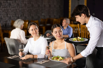 Amiable young waiter serving ordered dishes to carefree female friends gathered over dinner in restaurant