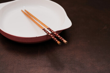 Empty white ceramic plate with  handle on the side and wooden chopsticks on the brown background, traditional wooden sticks for asian food.