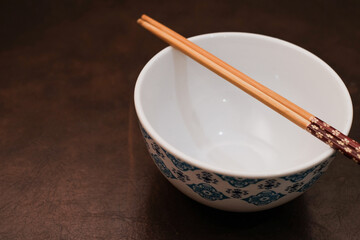 White bowl with blue pattern outside and wooden chopsticks on brown background, traditional wooden sticks for asian food.
