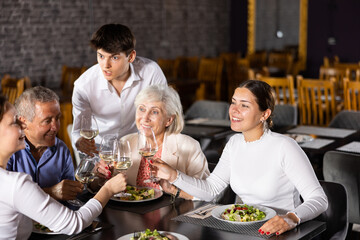 Cheerful elderly couple spending time with young friends in cozy restaurant. Man and woman having fun while talking and enjoying light dinner with wine at table