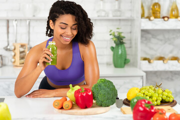 Vegetarian Food Concept. Portrait of smiling black girl drinking smoothie made of super foods, looking at vegetables on table