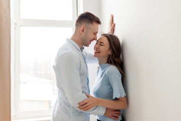 young happy couple in love stands near the window and laughs, the man hugs the woman and flirts at home and smiles