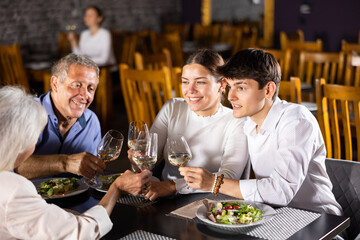Young couple enjoying delicious food and pleasant conversation with senior parents over dinner in restaurant. Happy carefree people gathered around table for family celebration..