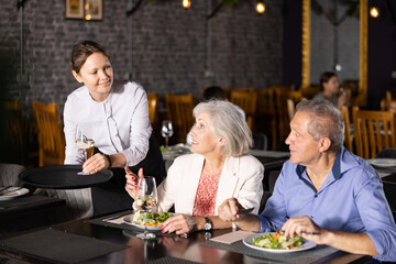 Hospitable smiling waitress serving ordered drinks to elderly couple sitting at table, visiting restaurant for romantic dinner