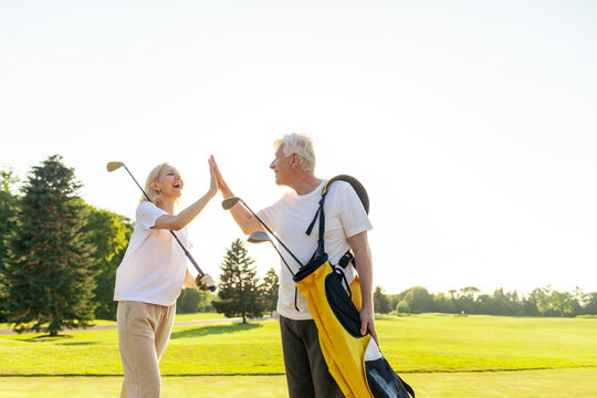 elderly senior couple in uniform celebrating victory and success in golf game and giving high five, old man and woman playing golf on golf course at sunset and doing outdoor sports