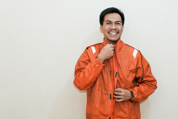 Smiling Southeast Asian man wearing orange raincoat with hands for zip up them looking at the camera. Positive person. standing over isolated white background. rainy season.