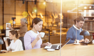 Positive European young woman drinking and enjoying coffee while working on laptop in cafe. Freelance and remote work