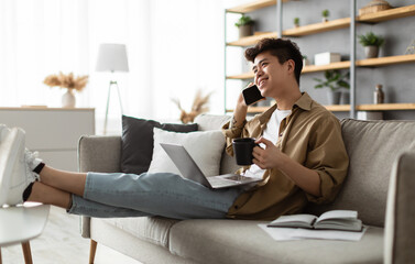 Mobile Communication. Portrait of smiling young asian guy using laptop and talking on mobile phone, holding cup and drinking coffee, working remotely sitting on the couch at home office