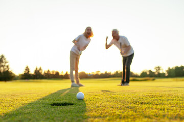 couple of elderly seniors in uniform playing golf on the course and celebrating victory at sunset, old man and woman hitting a golf ball into a hole and rejoicing in success together outdoors