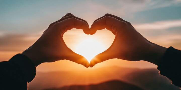 A close-up of hands forming a heart shape in front of a rising sun