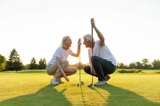 old man and woman in uniform playing golf on golf course at sunset, elderly couple having active outdoor recreation and doing sports, grandpa and grandma holding clubs and pointing where to hit the