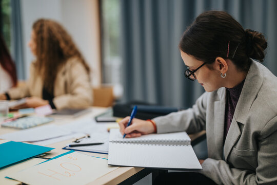 A student is attentively taking notes in a classroom setting, highlighting focus and learning. The environment is collaborative, with students dressed in business attire, creating a professional