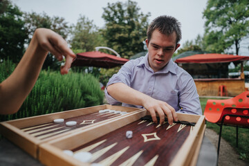 A boy with Down syndrome and a girl in a wheelchair share a joyful moment playing backgammon in an outdoor setting, emphasizing friendship and inclusion.