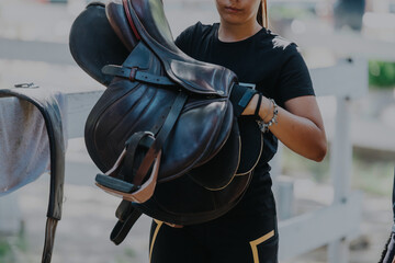 Close-up of a woman holding a horse saddle at an outdoor stable, ready for equestrian activities....