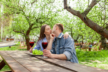 Naklejka premium young couple in love in denim clothes sitting in the garden at wooden table and drinking red wine with grapes on date and kissing in the summer, guy and girl trying alcoholic cocktails and flirting in