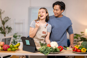 A man and woman are standing in a kitchen, smiling and eating vegetables