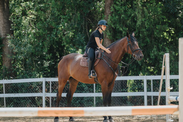 Young rider on horseback practicing riding skills in an outdoor equestrian arena surrounded by greenery and fencing.