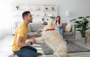 Happy young Arab guy playing with dog, having fun on floor at home, his girlfriend drinking coffee on background, copy space. Interracial married couple with cute pet spending time in living room