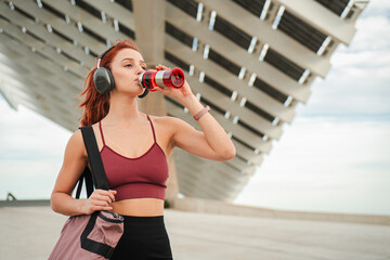 Young athletic woman in sports wear and headphones recovering drinking isotonic water from bottle...