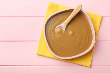 Delicious baby food and spoon in bowl on pink wooden table, top view