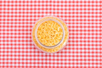 Uncooked Chifferi Rigati Pasta in Glass Jar on Checkered Red Towel on Pink Background. Fat and Unhealthy Food. Scattered Classic Dry Macaroni. Italian Culture and Cuisine. Raw Pasta