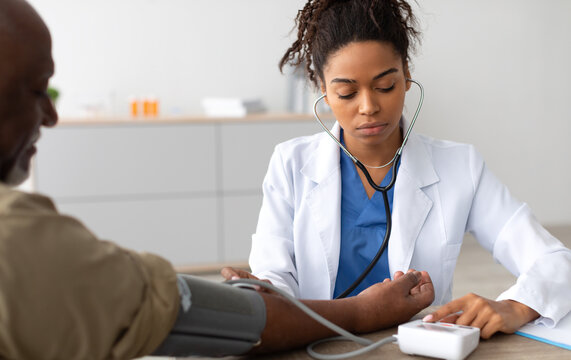 Young Focused African American Lady Examining Older Patient Measuring Arterial Blood Pressure With Stethoscope And Sphygmomanometer Medical Device Sitting At Table. Hypertension Health Problem Concept
