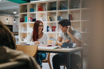 Two students sit together in a modern classroom, engaged in conversation and study. The atmosphere is relaxed and collaborative, with books and materials spread across the table.