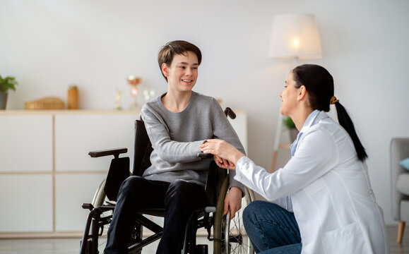 Disabled people healthcare support concept. Doctor holding hands of handicapped teen boy in wheelchair during medical visit. Happy impaired adolescent and young nurse at home