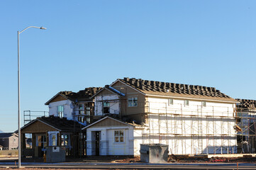 Two-story house under construction with exterior wrap, stacked roofing tiles, scaffolding, and adjacent utility structure, showing mid-phase residential development