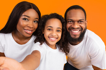 Photography Concept. Closeup portrait of smiling black family taking selfie picture, point of view pov shot of happy parents with daughter looking at camera making front selfportrait at yellow studio