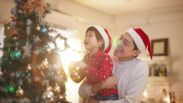 Happy family preparing New Year winter holidays celebration. Smiling young asian father and son enjoy decorating ornament on christmas tree create festive mood party atmosphere at home. slow motion. 
