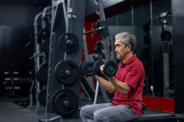 Naklejka premium Asian senior man in sportswear doing weight lifting on double dumbbells to gain bicep muscle while having exercise training in the gym for longevity and retirement