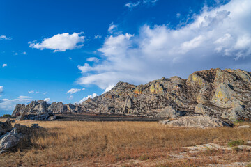 Dramatic layered sandstone cliffs rise above dry savanna grassland under bright blue sky with scattered white clouds. Jurassic geological formations at Isalo National Park, Madagascar.