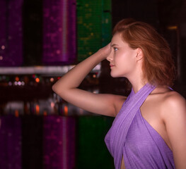 A young romantic girl against the background of modern buildings illuminated by multicolored LED light reflected in the water. Night city. Side view